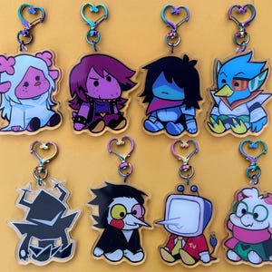Puede incluir: Colección de ocho llaveros acrílicos con personajes de dibujos animados. Cada llavero tiene un cierre en forma de corazón con un acabado arcoíris. Los personajes son de varios colores, incluyendo azul, morado y blanco, sobre un fondo amarillo.