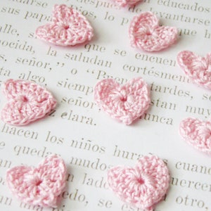 May include: A close-up of a page of text with several pink crocheted hearts scattered across the page. The hearts are all the same size and shape, and they are made with a delicate, lacy stitch.