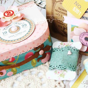 May include: A pink and white polka dot box with a teal and brown floral design. The box has a spool of thread with the text "Close Knit" and "Fine Thread" on it. The box is decorated with a pink flower and a number 29. The box is sitting on a white crocheted surface.