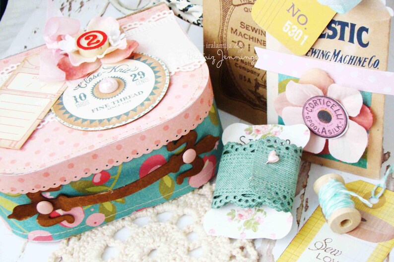 May include: A pink and white polka dot box with a teal and brown floral design. The box has a spool of thread with the text "Close Knit" and "Fine Thread" on it. The box is decorated with a pink flower and a number 29. The box is sitting on a white crocheted surface.