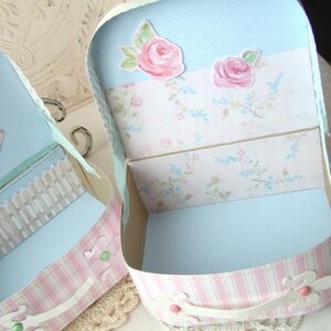 May include: A small, pink and white striped cardboard box with a blue and white floral patterned interior. The box has a hinged lid and is decorated with pink roses and a white lace trim.
