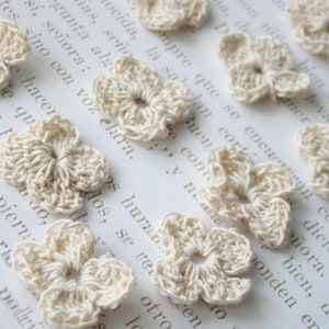 May include: A collection of ten off-white crocheted flower embellishments. The flowers are delicate and intricate, with a five-petal design.