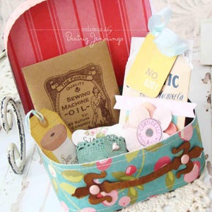 May include: A vintage-style floral fabric box with a brown handle and a metal clasp. The box is filled with various items, including a small sewing machine oil tin, a spool of thread, and several decorative tags. The text on the tags reads "No. 401", "Sewing Machine Co", and "The Finest Quality Sewing Machine Oil".
