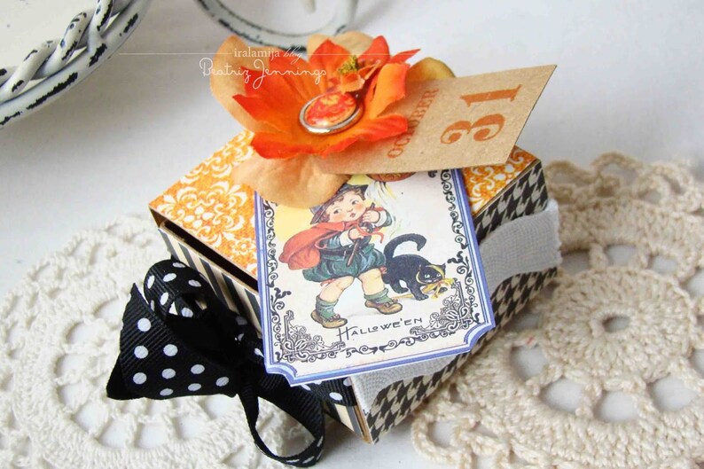 May include: A Halloween gift box with a black and white houndstooth pattern and a black polka dot bow. The box is decorated with an orange flower and a vintage Halloween card showing a child and a black cat. The tag reads "October 31".