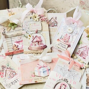 May include: A collection of handmade Christmas gift tags featuring pink and white paper, gingerbread houses, and festive text. The tags are decorated with ribbons, lace, and other embellishments.