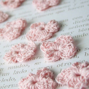 May include: A collection of small, light pink crocheted flower appliques. The flowers are arranged on a white surface with faint text visible in the background. The crochet work is detailed, with visible stitches.