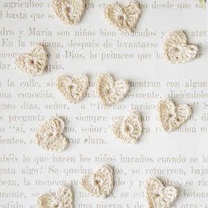 May include: A collection of small, crocheted hearts in a cream color. The hearts are arranged on a white surface with text in Spanish.