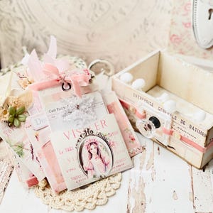 May include: A stack of vintage-style paper ephemera with pink and white accents, featuring a decorative box with a drawer and a small white knob. The paper ephemera includes a card with the text "VISER La TRES VIE 170 TOUTES A PLUS" and a card with the text "Cash Paid To".
