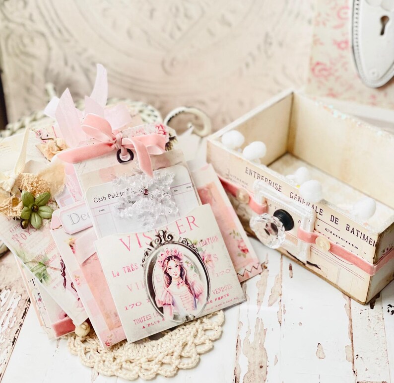 May include: A stack of vintage-style paper ephemera with pink and white accents, featuring a decorative box with a drawer and a small white knob. The paper ephemera includes a card with the text "VISER La TRES VIE 170 TOUTES A PLUS" and a card with the text "Cash Paid To".