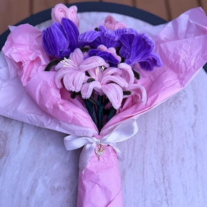 May include: A bouquet of handcrafted pipe cleaner flowers in shades of purple and light pink, wrapped in light pink tissue paper. The bouquet is tied with a white ribbon featuring a small, gold-toned ballerina charm with clear crystals.