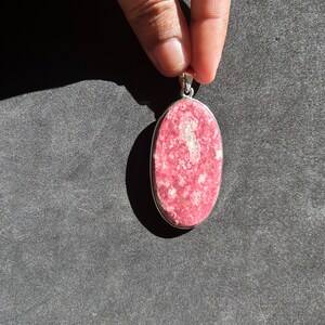 May include: A silver pendant featuring a pink and white oval gemstone. The pendant is displayed against a black backdrop.