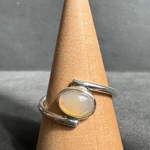 May include: A silver ring featuring an oval opal gemstone. The ring's band wraps around the finger, with the opal set in a bezel. The opal displays a milky white base with touches of yellow and orange.
