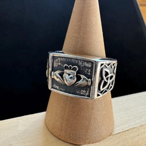 May include: A silver-toned ring featuring a Claddagh design with a heart, crown, and hands. The ring has a rectangular shape with Celtic knot detailing on the sides. The ring is displayed on a brown, cone-shaped stand.