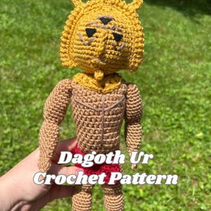 Peut inclure: Poupée au crochet faite à la main avec une tête jaune et un corps marron, portant un short rouge. La poupée a trois yeux noirs et un couvre-chef en forme de couronne. Le texte "Dagoth Ur Crochet Pattern" est affiché sur la poupée.