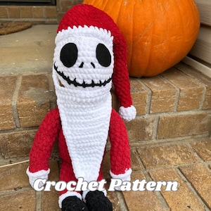 Op de afbeelding: Een handgemaakte gehaakte pop in de gelijkenis van Jack Skellington, met een rode kerstmuts en pak met witte details. De pop heeft een wit gezicht met zwarte ogen en een gestikte glimlach. De tekst "Crochet Pattern" is zichtbaar. Een oranje pompoen staat op de achtergrond.