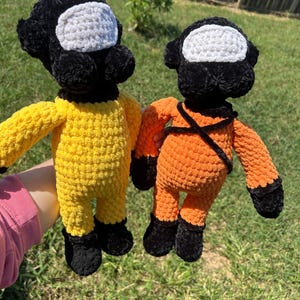 May include: Two handmade crochet dolls, one yellow and one orange, resembling characters from a popular video game. Each doll has a black head with a white visor, black feet, and matching black accents. The orange doll has black straps.