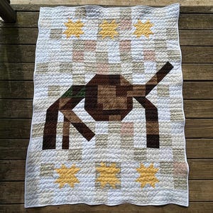 May include: A rectangular quilt featuring a central brown geometric elephant figure against a background of white, beige, and light pink squares. The design includes yellow star-shaped accents in the corners and a white border with wavy stitching.