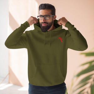 Puede incluir: Sudadera con capucha verde oliva con bolsillo de canguro y un gráfico rojo en la parte superior izquierda del pecho. La sudadera tiene una capucha con cordón y mangas largas. La persona de la imagen lleva gafas y tiene barba.