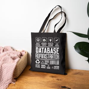 Puede incluir: Una bolsa de tela negra con texto e iconos gráficos en blanco. El diseño incluye las palabras DATABASE ADMINISTRATOR, junto con iconos de multitarea, café, resolución de problemas y viajes, además de etiquetas de advertencia y un código de barras.