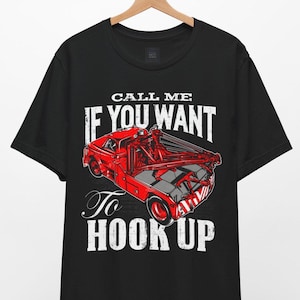 Può includere: Una t-shirt nera a maniche corte con la grafica di un carro attrezzi rosso. Il testo "CALL ME IF YOU WANT To HOOK UP" è stampato in lettere bianche dall'aspetto usurato sopra e sotto l'illustrazione del camion, che include braccio e verricello.