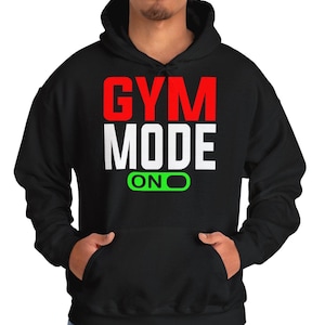 Puede incluir: Sudadera con capucha negra con el texto "GYM MODE ON" en letras blancas y rojas. La palabra "GYM" está en rojo, y el resto del texto está en blanco. Un gráfico de interruptor de luz verde está debajo del texto.
