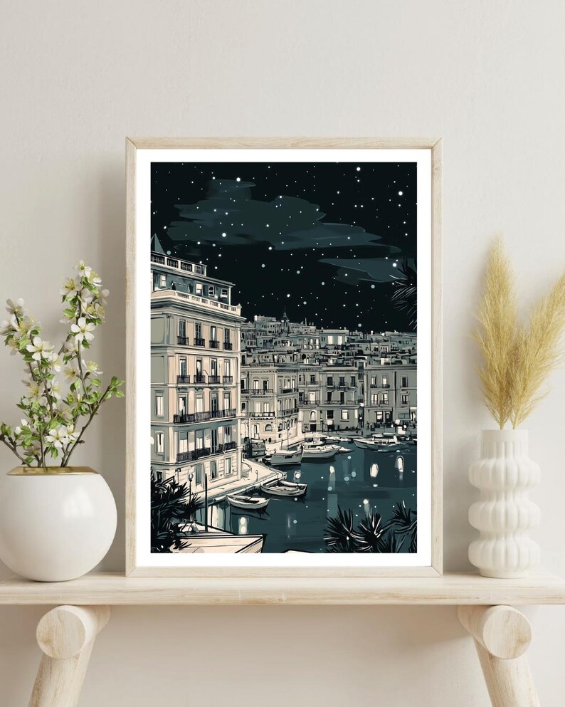 May include: A framed art print depicting a nighttime harbor scene. The illustration features buildings with lit windows, boats on the water, and a starry sky. The color palette includes shades of blue, white, and gray.