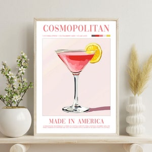 May include: A framed poster with a pink Cosmopolitan cocktail in a martini glass with a lemon wedge garnish. The poster has the text "COSMOPOLITAN" and "MADE IN AMERICA" with the recipe for the drink listed below.