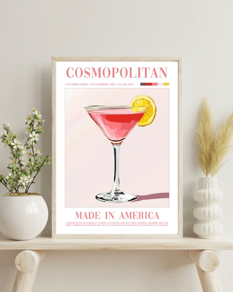 May include: A framed poster with a pink Cosmopolitan cocktail in a martini glass with a lemon wedge garnish. The poster has the text "COSMOPOLITAN" and "MADE IN AMERICA" with the recipe for the drink listed below.