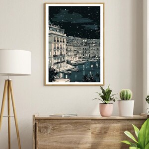 May include: A framed art print depicts a nighttime harbor scene. Buildings with lit windows line the water's edge, with boats docked below. The sky is dark with stars and clouds. The print is in a gold frame and is displayed on a wooden cabinet.