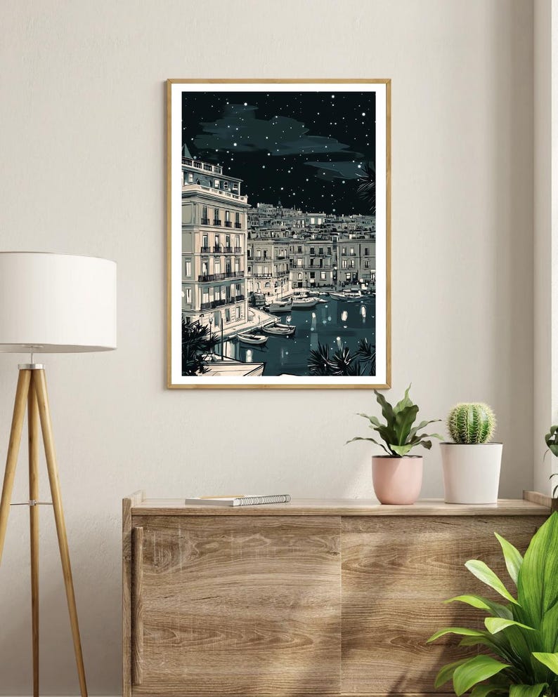 May include: A framed art print depicts a nighttime harbor scene. Buildings with lit windows line the water's edge, with boats docked below. The sky is dark with stars and clouds. The print is in a gold frame and is displayed on a wooden cabinet.