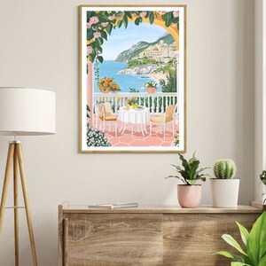 May include: A framed illustration of a pink and white patio with a table and chairs overlooking a blue ocean and a town on a hillside. The patio is covered in a yellow and green vine with pink flowers.