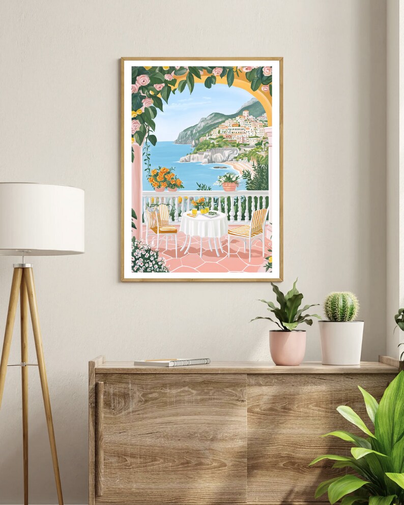 May include: A framed illustration of a pink and white patio with a table and chairs overlooking a blue ocean and a town on a hillside. The patio is covered in a yellow and green vine with pink flowers.