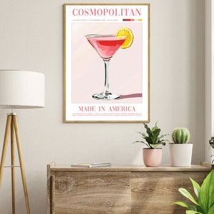May include: A pink Cosmopolitan cocktail with a lemon wedge garnish. The drink is in a martini glass and the text "COSMOPOLITAN" and "MADE IN AMERICA" is on the poster. The ingredients are listed as 1.5 oz vodka, 1 oz cranberry juice, and 1/2 oz lime juice.