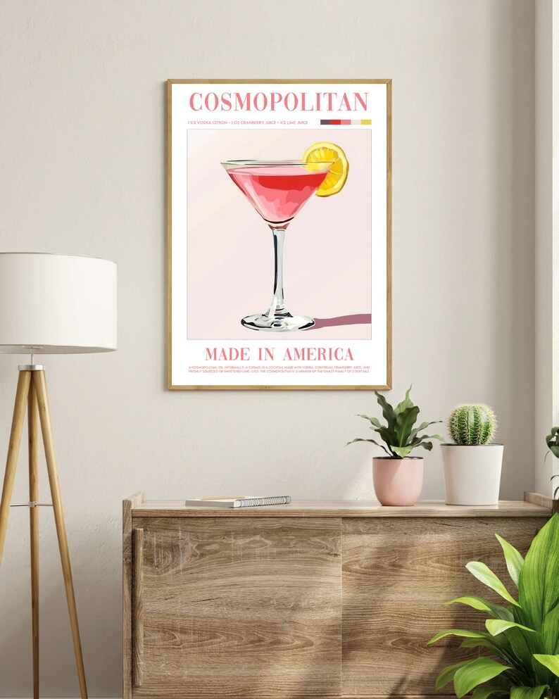 May include: A pink Cosmopolitan cocktail with a lemon wedge garnish. The drink is in a martini glass and the text "COSMOPOLITAN" and "MADE IN AMERICA" is on the poster. The ingredients are listed as 1.5 oz vodka, 1 oz cranberry juice, and 1/2 oz lime juice.