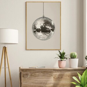 May include: A silver disco ball hanging from the ceiling against a beige background. The disco ball is made of many small mirrors and is reflecting light.