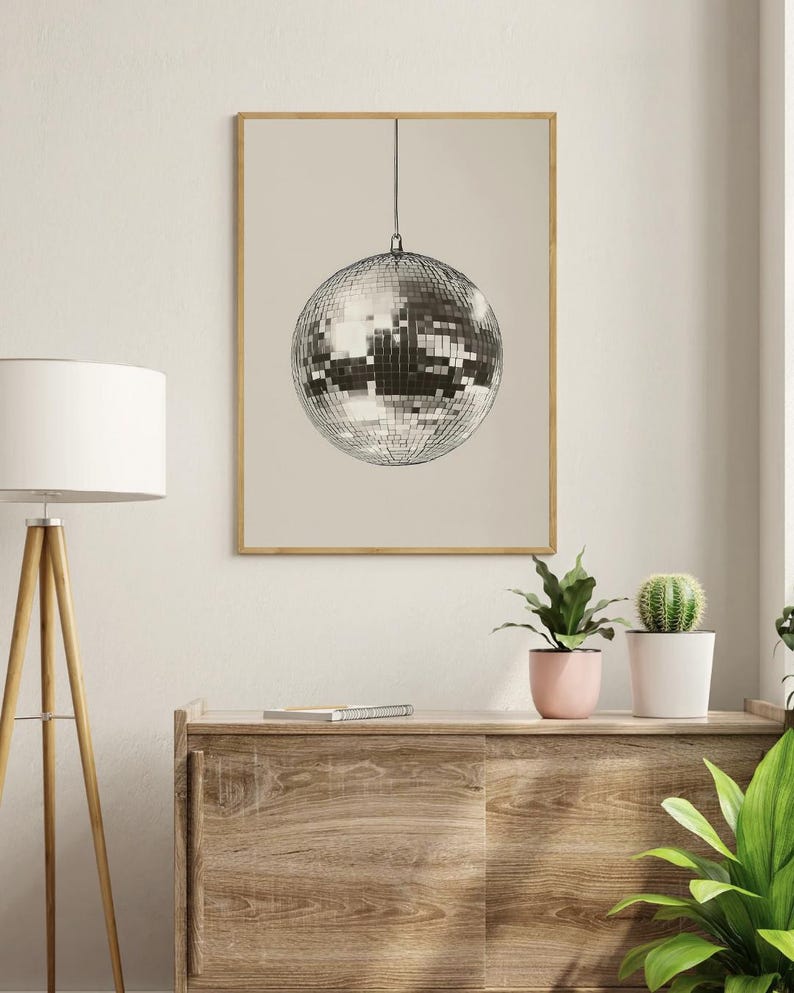May include: A silver disco ball hanging from the ceiling against a beige background. The disco ball is made of many small mirrors and is reflecting light.