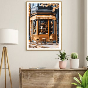 Op de afbeelding: Een schilderij van een winkelgevel met een gele luifel en twee stoelen en een tafel ervoor. De winkelgevel heeft een groot raam met een etalage van goederen erin. Het gebouw is geschilderd in tinten blauw en geel.