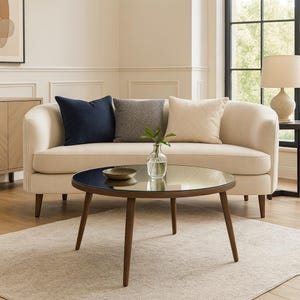 May include: A living room scene featuring a cream-colored sofa with three decorative pillows in navy blue, gray, and cream. A round coffee table with a dark glass top and wooden legs sits in front of the sofa. A small vase with greenery is on the table.