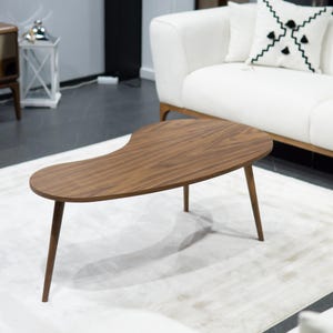 Op de afbeelding: Een salontafel in mid-century modern stijl met een niervorm. De tafel is gemaakt van donkerbruin hout en heeft vier taps toelopende poten. De tafel staat op een wit tapijt voor een witte bank.