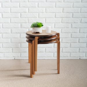 May include: A set of three nesting tables with a light brown finish. The tables have round tops and are stacked together. A small white ceramic pot with a green plant and a white candle sit on top of the tables.