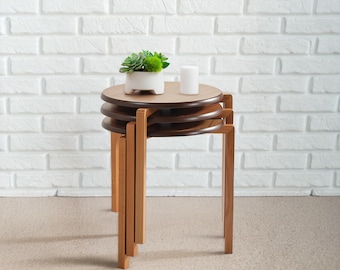 May include: A set of three stacked round wooden stools with dark brown circular tops and light brown legs. A white ceramic planter containing a green succulent plant and a small white cylindrical candle sit on the top surface of the stool set.