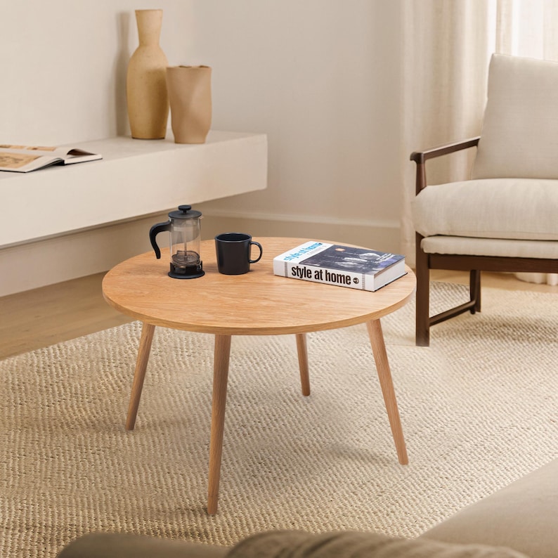 Pu&ograve; includere: Un tavolino da caff&egrave; rotondo in legno con quattro gambe di colore marrone chiaro. Sul tavolo ci sono una tazza da caff&egrave; nera, una french press di vetro e un libro intitolato "Style at Home".