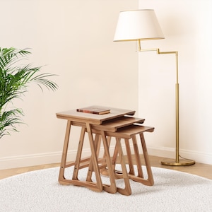May include: Three wooden nesting tables in a natural finish, arranged in a stacked formation. A small book rests on the top table. The tables are set on a white rug, with a gold floor lamp and a green plant visible in the background. The tables are approximately 60cm in height.