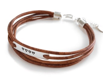 May include: A brown leather bracelet with a silver charm that has four small hearts engraved on it.