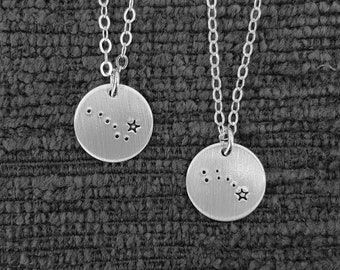 May include: Two silver necklaces with matching round pendants featuring a constellation design with small stars. The pendants are on a textured gray background.