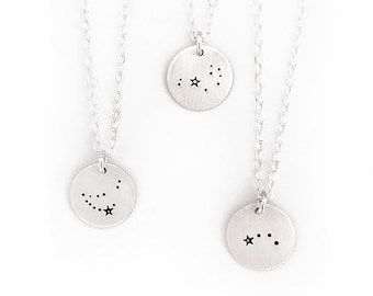 May include: Three silver-toned necklaces with round pendants, each featuring a constellation design. The pendants are suspended from delicate chains. The background is white, and the necklaces are arranged in a vertical composition.