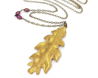 May include: A gold oak leaf pendant necklace with a chain and two small purple beads. The necklace is on a white background.