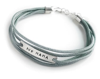 May include: A gray multi-strand bracelet with a silver bar charm that reads "IVF MAMA".