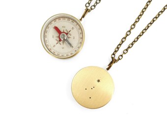May include: A gold necklace with two pendants. The first pendant is a compass with the words "Stanley London" printed on it. The second pendant is a round disc with a small star and four dots arranged in a constellation pattern.