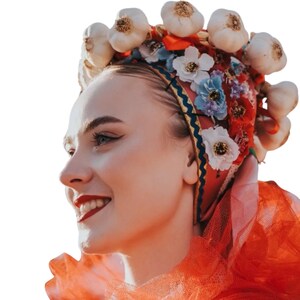 Puede incluir: Una mujer con un tocado tradicional con bulbos de ajo, flores y una bufanda roja. El tocado presenta detalles blancos, azules y rojos. La mujer sonr&iacute;e, con l&aacute;piz labial rojo y delineador de ojos oscuro.